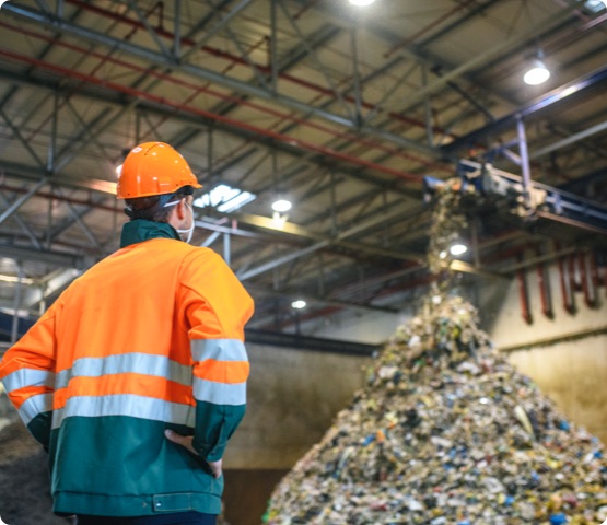 Low angle rear view of young male worker in helmet, pollution mask, and reflective clothing observing waste falling from conveyor belt onto pile at facility.