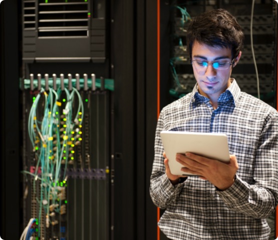 Professional IT programming technician is in a server room, checking the main computer equipment. The man has short brown hair, and he is wearing glasses and a casual black-and-white plaid shirt. A Digital Tablet is attached to the computer server amid colorful blue computer cables and wires.